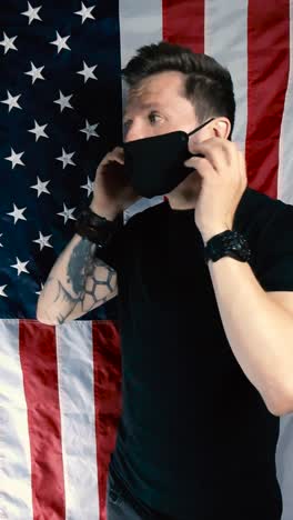 tattooed man in front of a vertical hanging american united states flag puts on a black colored health medical protection mask the right way appropriately and points at it while nodding with approval