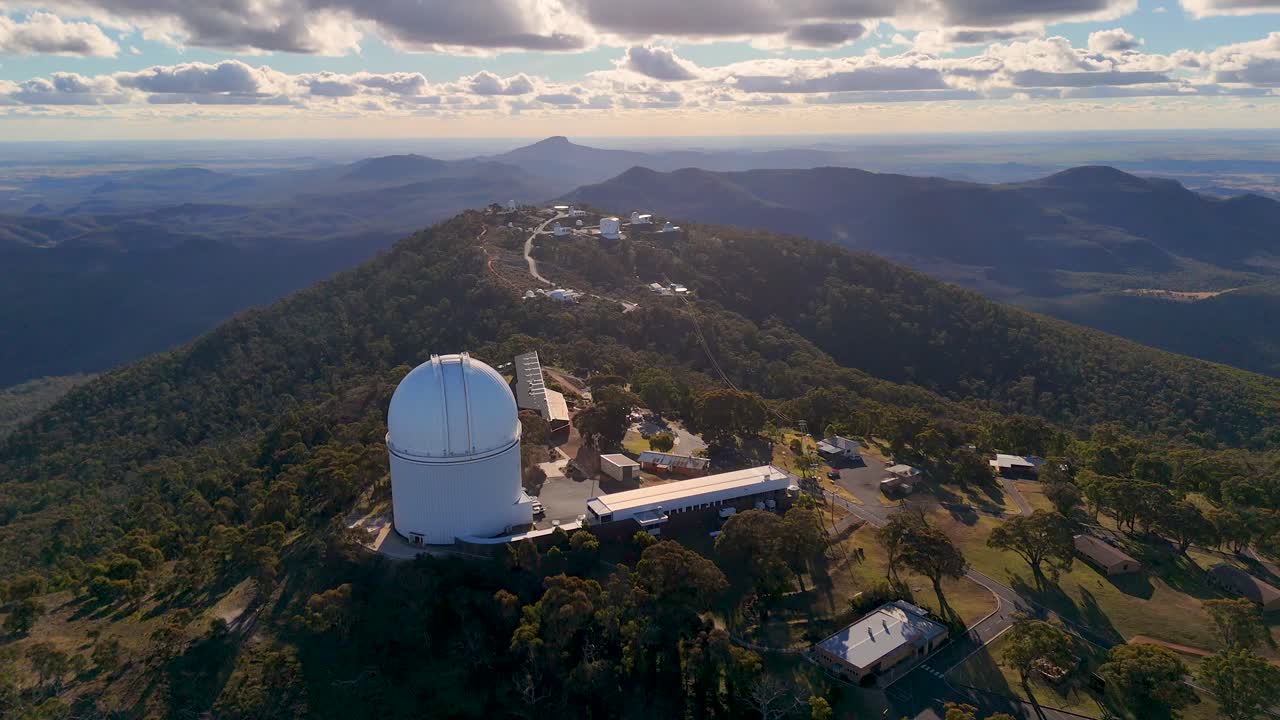 Drone camera glides over a large observatory dome atop a forested mountain ridge, revealing expansive sunset views and dramatic cloud formations
