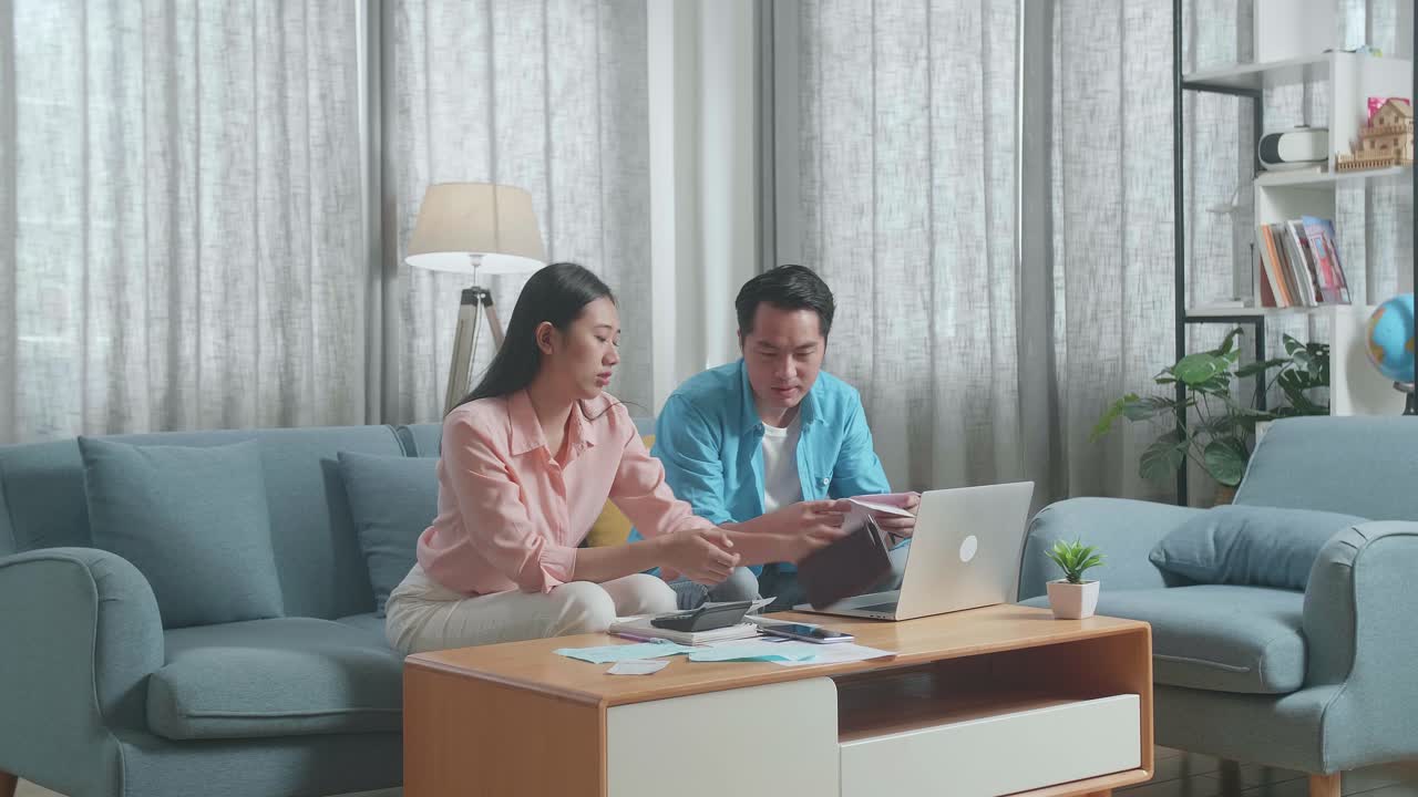 アジアのカップルがノートパソコンを持って請求書を見て財布の金をチェックしその後頭を振り頭痛を感じています
