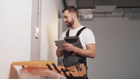 portrait of a man in a company work uniform with a tool belt and tablet in hand, configuring an access panel for controlling ventilation, air conditioning, security alarm, and surveillance cameras