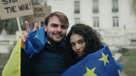 embraced caucasian man and woman covered with eu and ukraine flags are showing a peace sign.