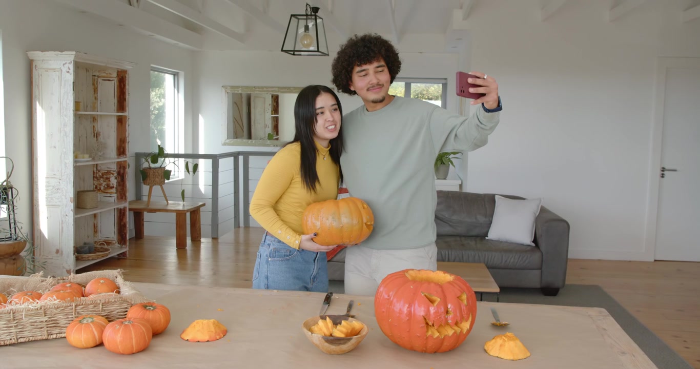 Coppia consulta lo smartphone e prepara una zucca per intagliarla ad Halloween, mettendosi in posa per un selfie a casa