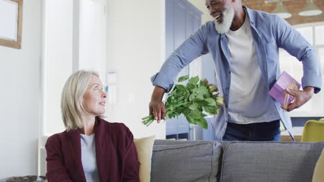 happy senior diverse couple in living room sitting on sofa, giving flowers and present