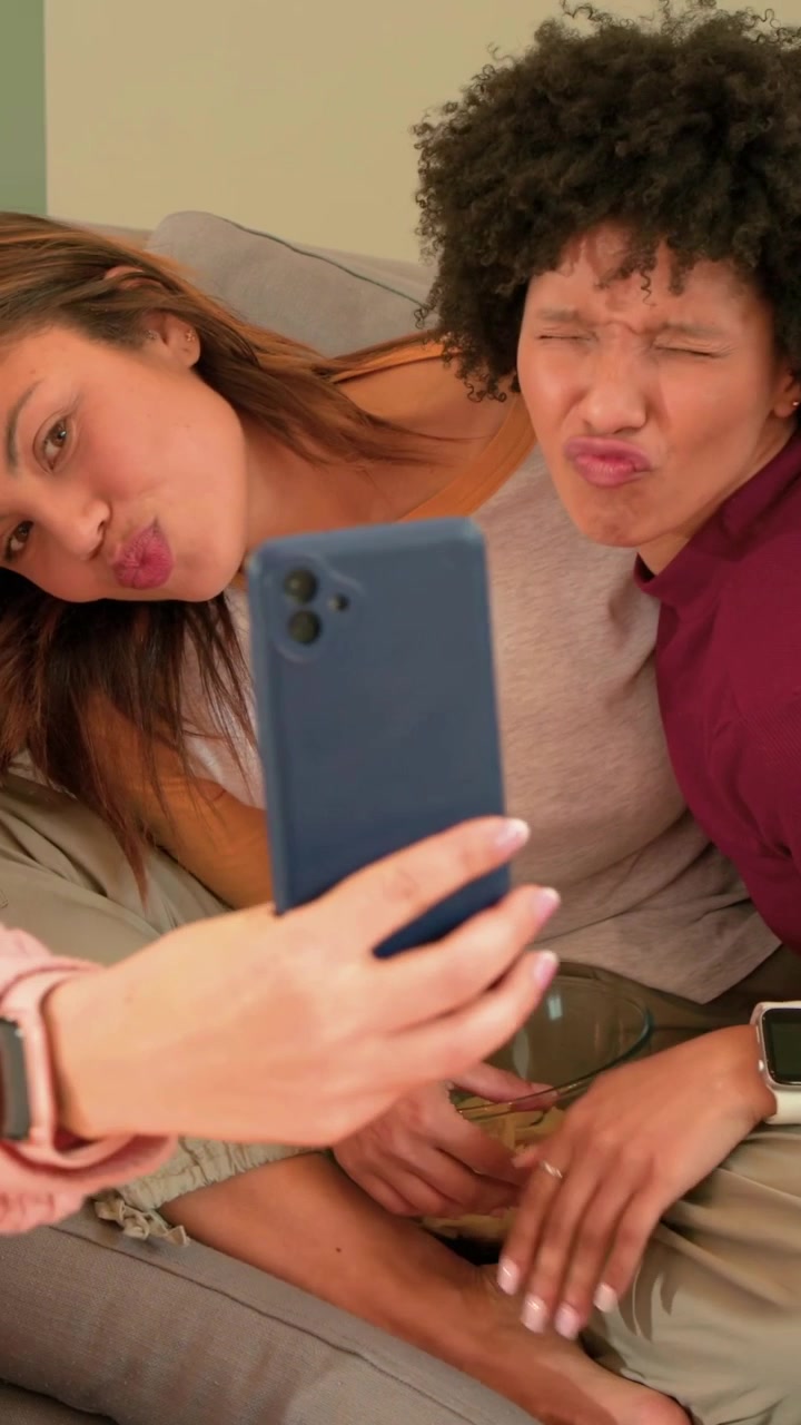 Vertical video: Left raising blue phone, women maroon and raglan leaning on couch, taking selfie