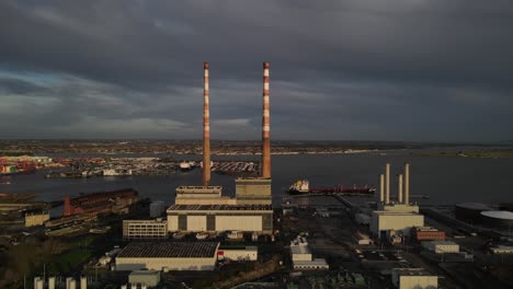 Chimeneas-Antiguas-De-La-Estación-Generadora-Poolbeg-En-Dublín,-Irlanda-Bajo-Un-Cielo-Oscuro-Con-El-Puerto-De-Dublín-En-El-Fondo---Amplia-Toma-Aérea