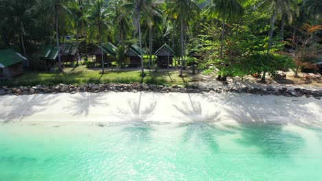 cabañas de playa bajo palmeras con sombra de hojas verdes sobre arena blanca de playa exótica bañada por una tranquila laguna turquesa