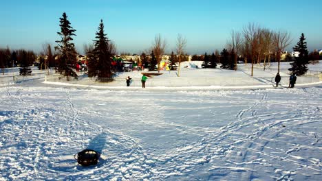 Winteraufstieg-Aus-Der-Luft-Fliegen-Sie-über-Schneebedeckte-Heuhaufen-Picknicktische-Neben-Eisernen-Feuerstellen-Mit-Optionalen-Grills-Rund-Um-Eine-Künstliche-Eisbahn,-Die-Mit-Einer-Eisbahn-Um-Einen-Spielplatz-Verbunden-Ist-1-2