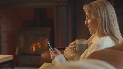 woman at home in lounge with cosy fire and hot drink using mobile phone