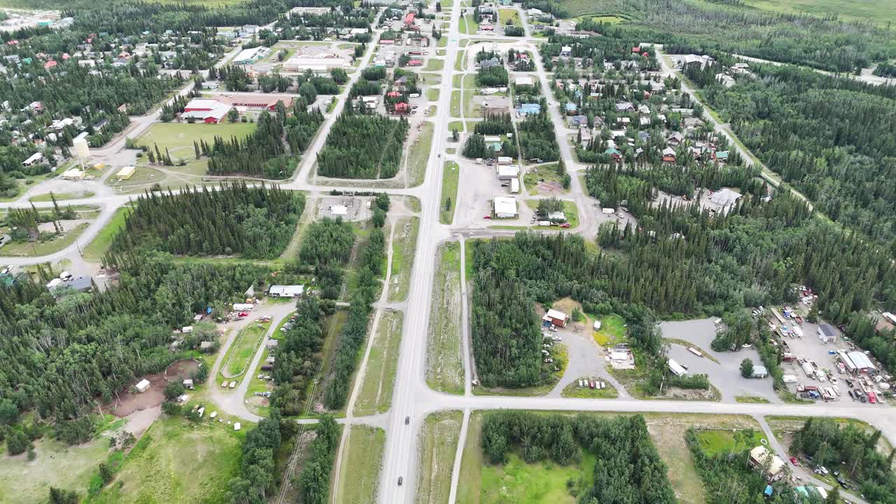 Drone perspective captures a remote northern town with dense forest surrounding residential streets and a central highway in Alaska, showcasing community life