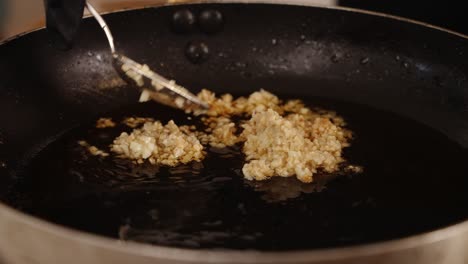 chef usando una cuchara para esparcir el ajo picado, asegurándose de que esté cubierto uniformemente con el aceite de cocina, friendo lentamente el ajo en la sartén poco profunda a fuego lento, primer plano