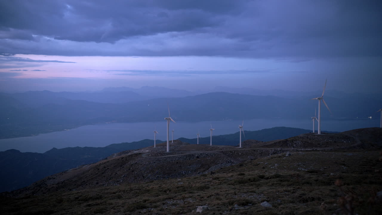 그리스 해안 계곡을 내려다보는 산꼭대기 위의 풍력 터빈, 황혼의 푸른 하늘 아래에서 낮에서 밤으로 전환하는 동안
