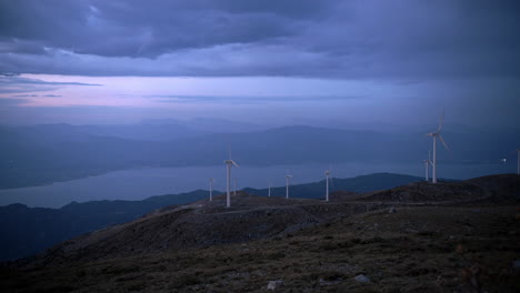turbinas eólicas en una cresta de montaña con vistas a un valle costero griego al anochecer, durante la transición del día a la noche bajo un cielo azul temperamental
