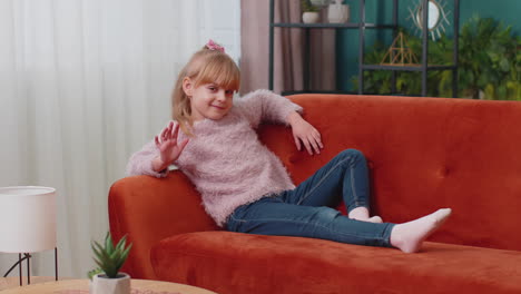 niña acostada en el sofá en casa mirando a la cámara, sonriendo, agitando las manos haciendo gestos de saludo o adiós