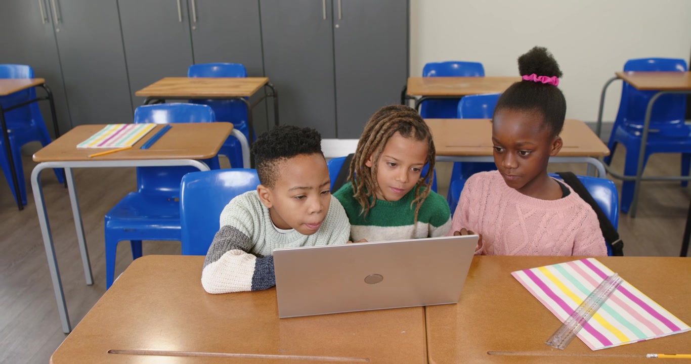 Diversi studenti in età scolare chinati verso un laptop aperto, che digitano e usano il touchpad per un compito.