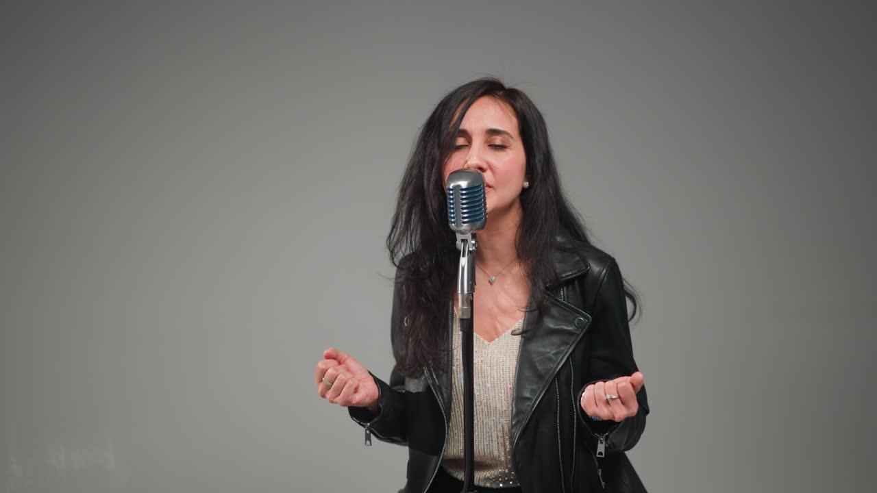 Emotional female singer in a leather jacket and sequined top performing at a vintage microphone against a white background