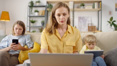 female teacher having online class at home and showing charts and statistics while her children sitting on sofa behind her using electronic devices