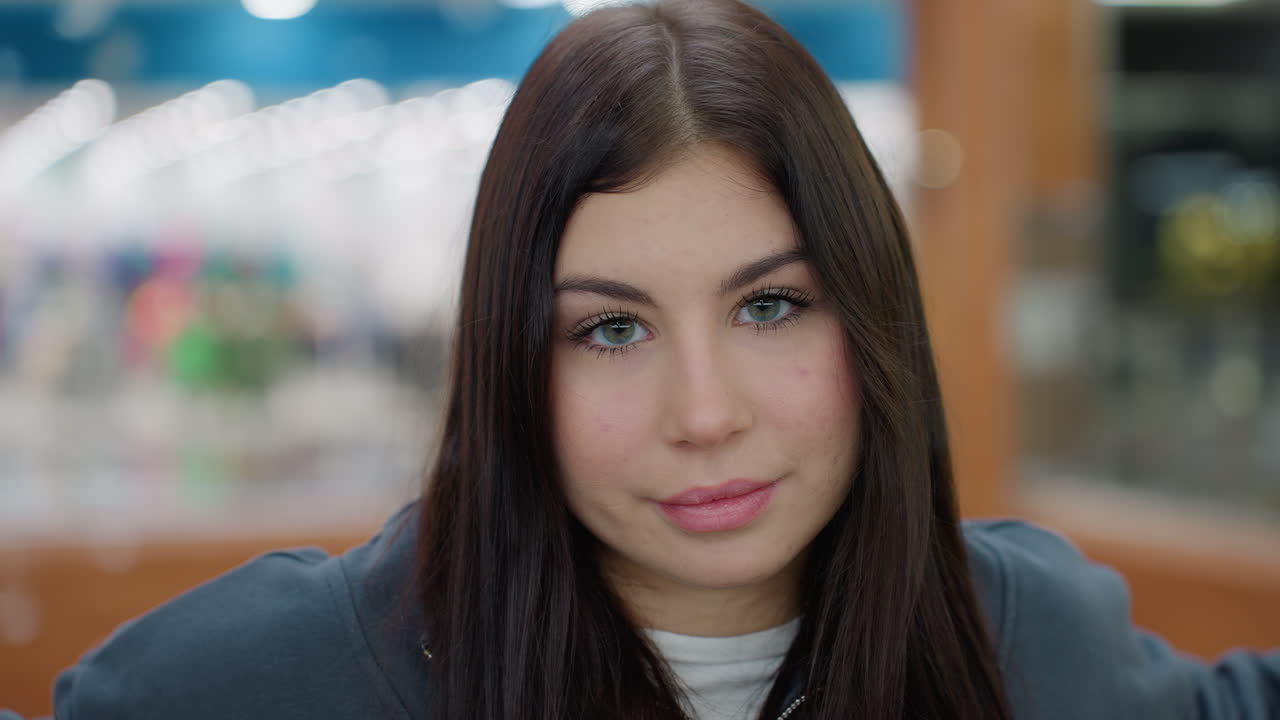 Portrait of beautiful young lady with long hair in dark sweatshirt staring calmly into camera with relaxed face and arms resting wide inside bright modern shopping mall with blurred lights and people
