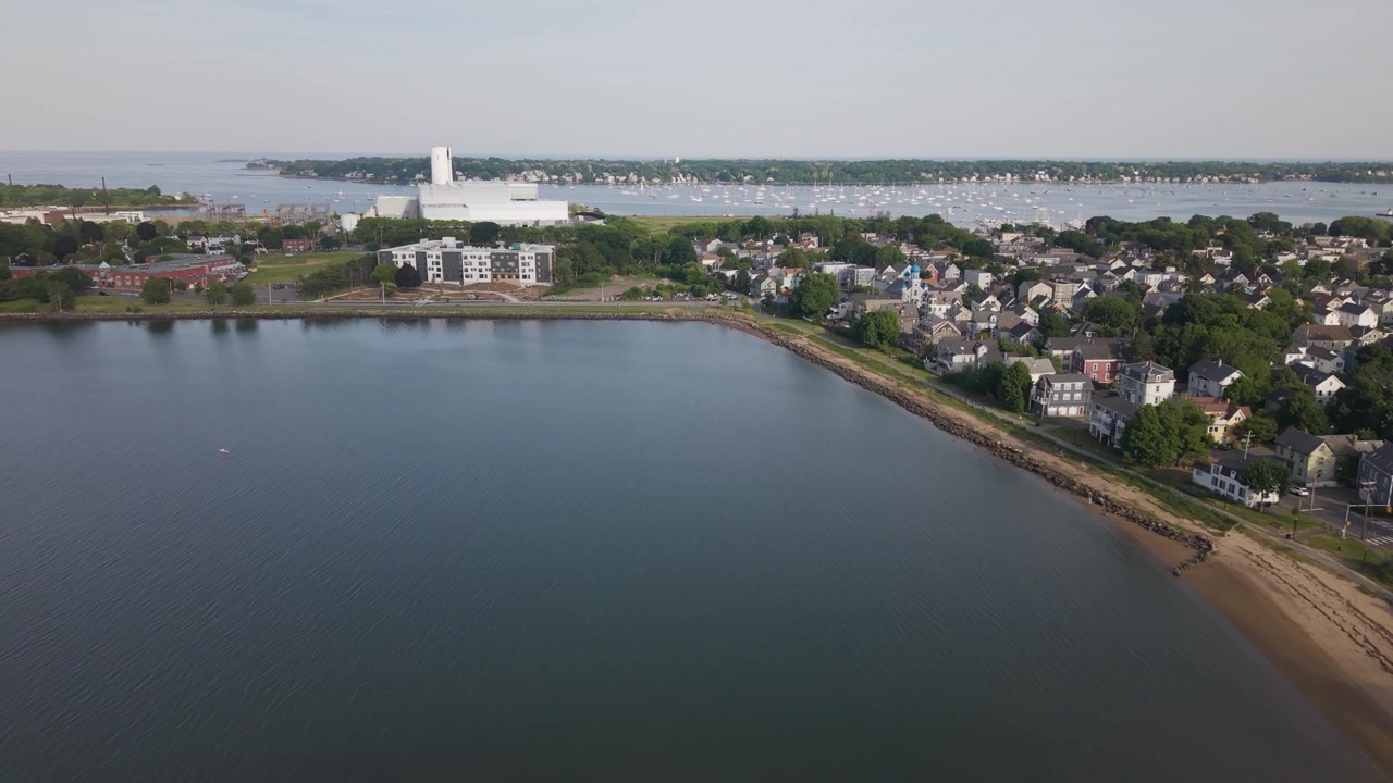 Una laguna calma si snoda oltre un campo da baseball sabbioso e un campo esterno verde, un denso quartiere costiero si estende verso il porto pieno di barche a vela a Salem, Massachusetts, USA. Ripresa aerea con drone a volo d'uccello
