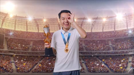 hombre asiático con una medalla de oro y un trofeo mostrando la mano de acuerdo firma sobre el ojo y sonriendo a la cámara como el primer ganador en el estadio