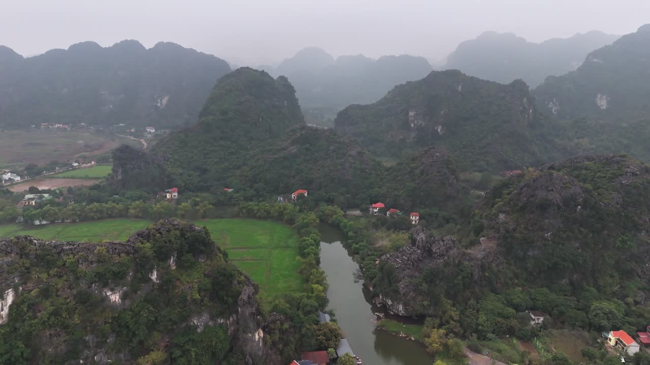 Aerial view of Trang An in Ninh Binh, Vietnam, featuring a small village nestled between towering limestone mountains and winding waterways under an overcast sky