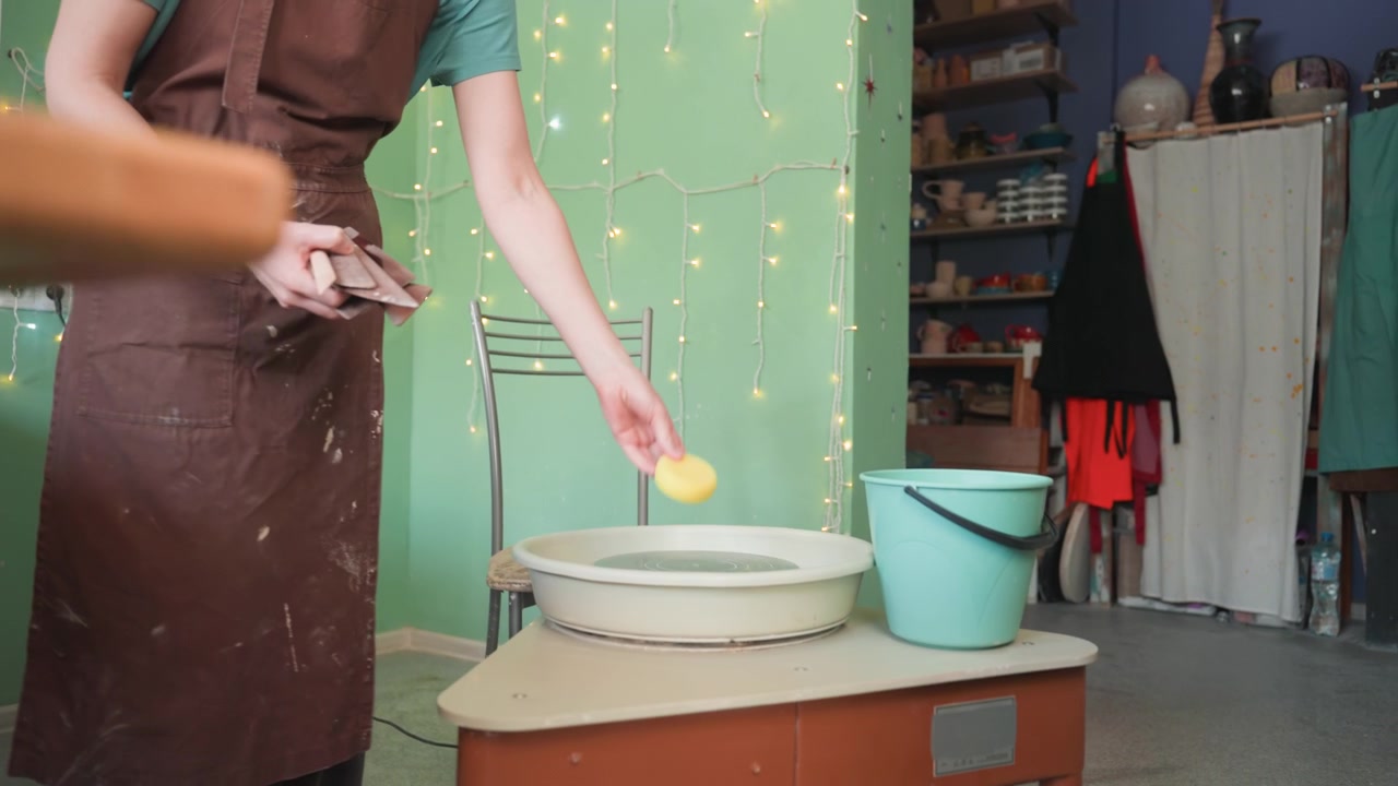 Giovane donna bianca che modella l'argilla al tornio, con un grembiule marrone, in un accogliente laboratorio di ceramica con lucine e scaffali, mani che levigano l'argilla umida, secchio e spugna nelle vicinanze, che mostra una lezione artigianale focalizzata
