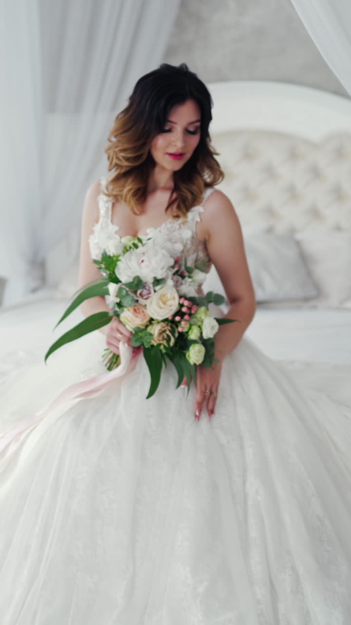 Attractive young bride in a sexy wedding dress is sitting on bed. Beautiful bride with flowers. Pretty model in white dress is posing to the female photographer on the bedroom background. Vertical video