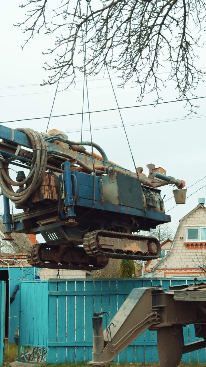 Special machine for well boring hanging on the crane hook. Rural village at backdrop. Vertical video.