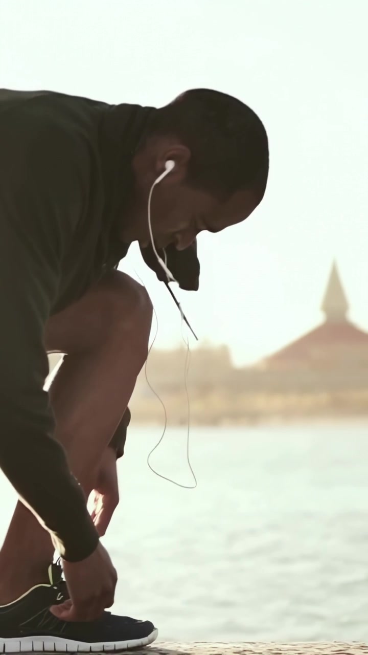 Vertical video: Walking hoodie man stopping to fix loose lace and earbuds at pier, copy space