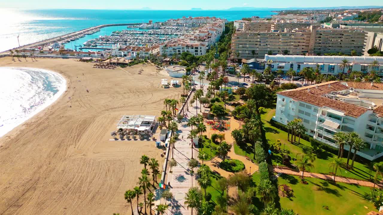 Ripresa aerea inclinata verso l’alto della spiaggia e del porto di Puerto Banús, Marbella, Costa del Sol, Spagna, con edifici di lusso color terracotta, lungomare alberato con palme e pedoni, barche nel porticciolo e la statua de La Victoria