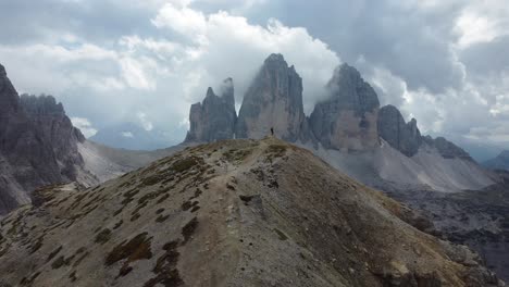 a slow-moving drone shot flying away from a hiker standing alone in the mountains of the dolomites in south tyrol in italy - he is looking at the outstanding rock formations of tre cime di lavaredo