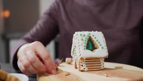 woman reaching to pick up golden bead rolling on wooden tray while decorating gingerbread house, intricate icing design with green beads, dried flowers, and blurred candlelight in background