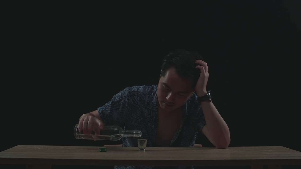 Drunk, Depressed Asian Man Pouring Vodka In A Shot Glass Before Drinking In Black Background