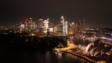 forward drone flight reveals sydney’s glittering night skyline, sweeping over circular quay toward the opera house and harbour bridge while bright reflections ripple across the dark harbour waters.