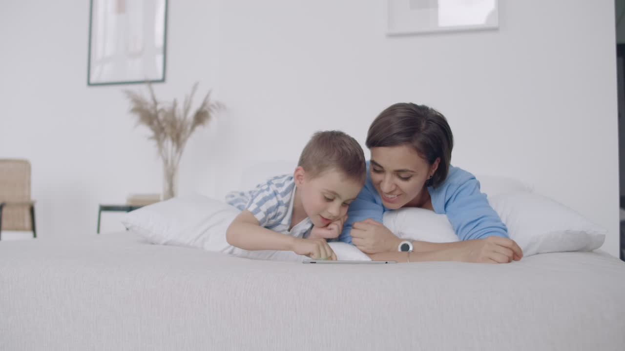 mamá e hijo mirando la pantalla de la tableta tendida en una cama blanca. juega juegos con tu hijo en tu tableta y mira videos divertidos.