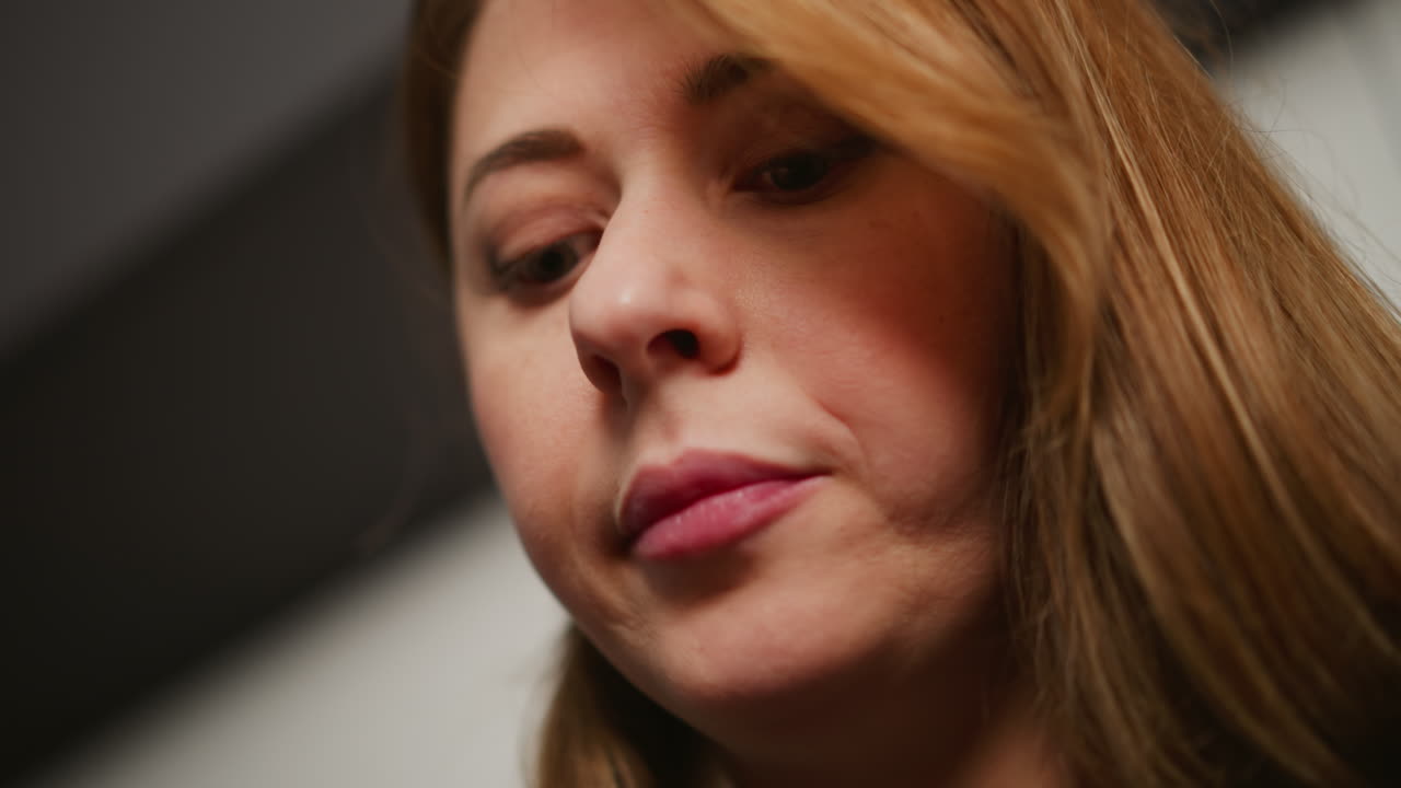 Extreme close up of woman with full flowing hair chewing food thoughtfully in kitchen. Subtle expression shows casual enjoyment of meal under soft indoor lighting