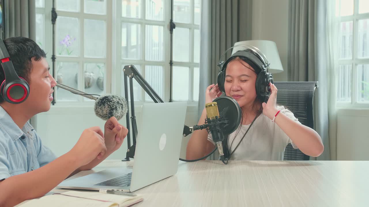 Asian Kid Boy In Headphones Singing Into Microphone While Recording Podcast With Boy Host In Studio
