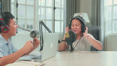 asian kid boy in headphones singing into microphone while recording podcast with boy host in studio