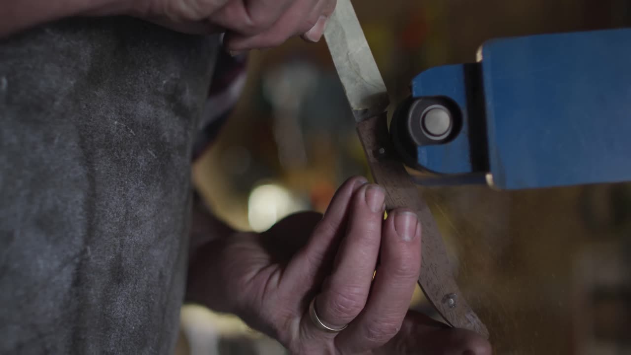 primer plano de un hombre caucásico fabricante de cuchillos en un taller sosteniendo un cuchillo y usando una lijadora