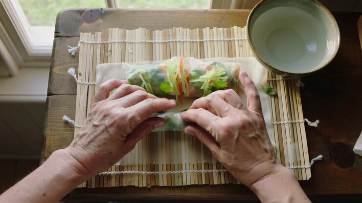 Posizionamento di erbe aromatiche, mani a maniche corte che piegano e arrotolano carta di riso su un tappetino di bambù vicino alla finestra, con una ciotola