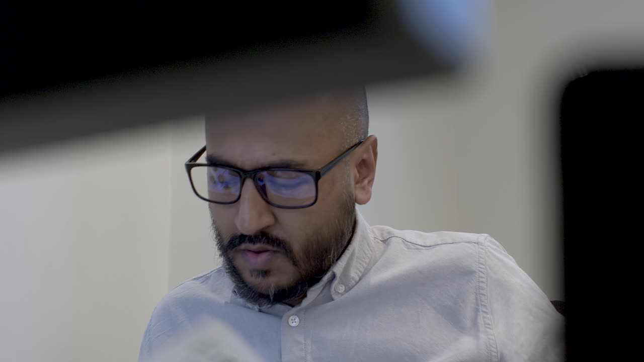 Imprenditore calvo di minoranza etnica che si volta per leggere alla scrivania, visto da dietro i monitor del computer.