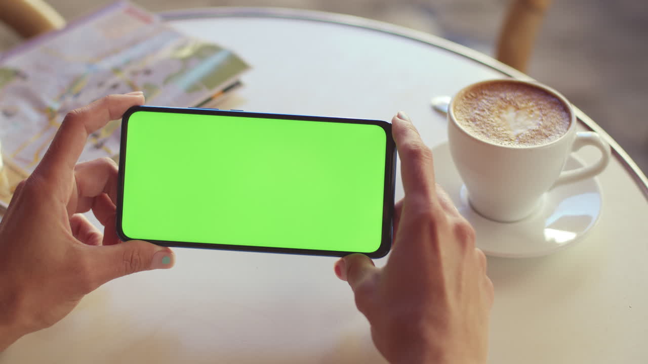 Hands holding a smartphone with a green screen at a cafe table with coffee