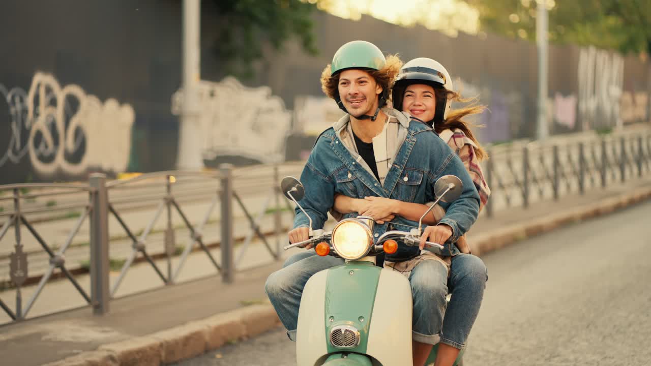 Happy couple, a guy with long curly hair in a denim jacket rides with his happy girlfriend in a plaid shirt who looks out from behind him on a green moped with the lights on along a street with a small fence or barrier in a summer city
