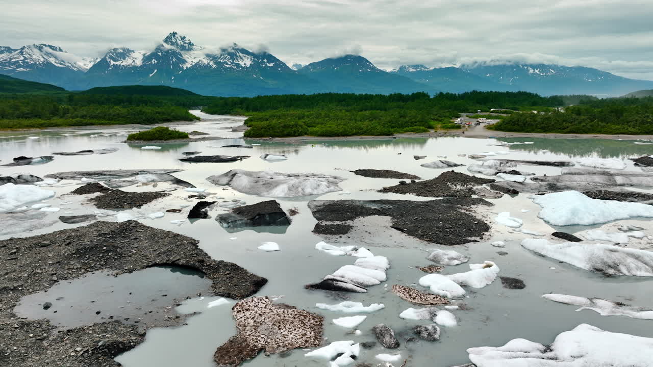 Pieces of melting ice in the river. Green waterfront and beautiful mountains at backdrop. Alaska wilderness