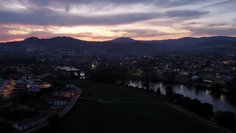 Dusk-settles-on-Barcelos,-ancient-bridge-and-reflective-river-stand-out-against-the-softly-lit-Portuguese-town---aerial