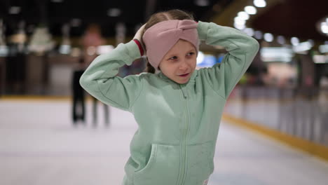 a close view of a child in a mint green outfit and pink headband dancing joyfully on an ice rink, the background shows other people skating and watching