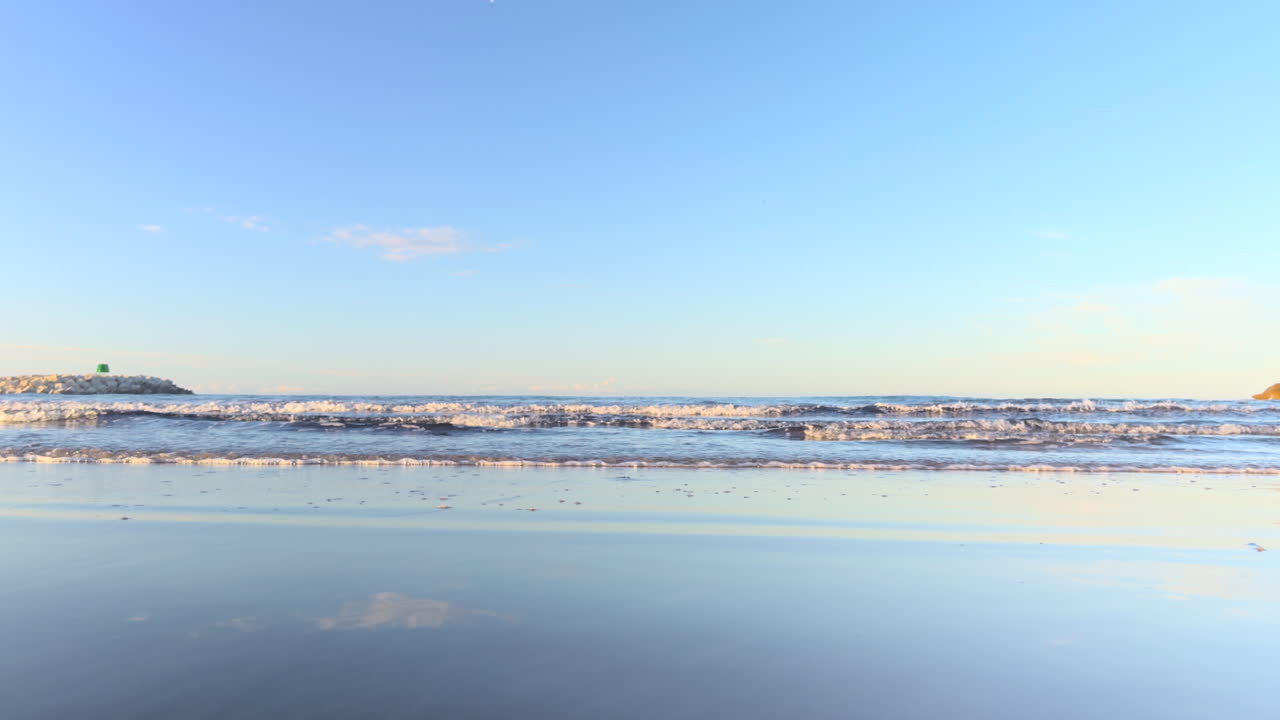 Low-angle shot of gentle waves breaking on the shore with sky reflections on the wet sand. Calm seascape during daylight under a clear blue sky