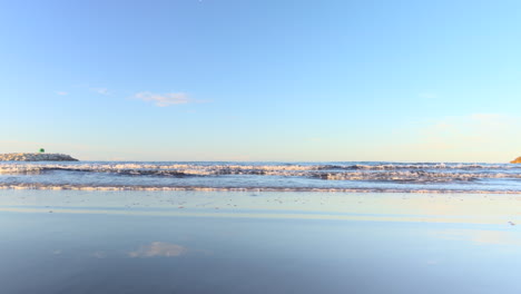 low-angle shot of gentle waves breaking on the shore with sky reflections on the wet sand. calm seascape during daylight under a clear blue sky