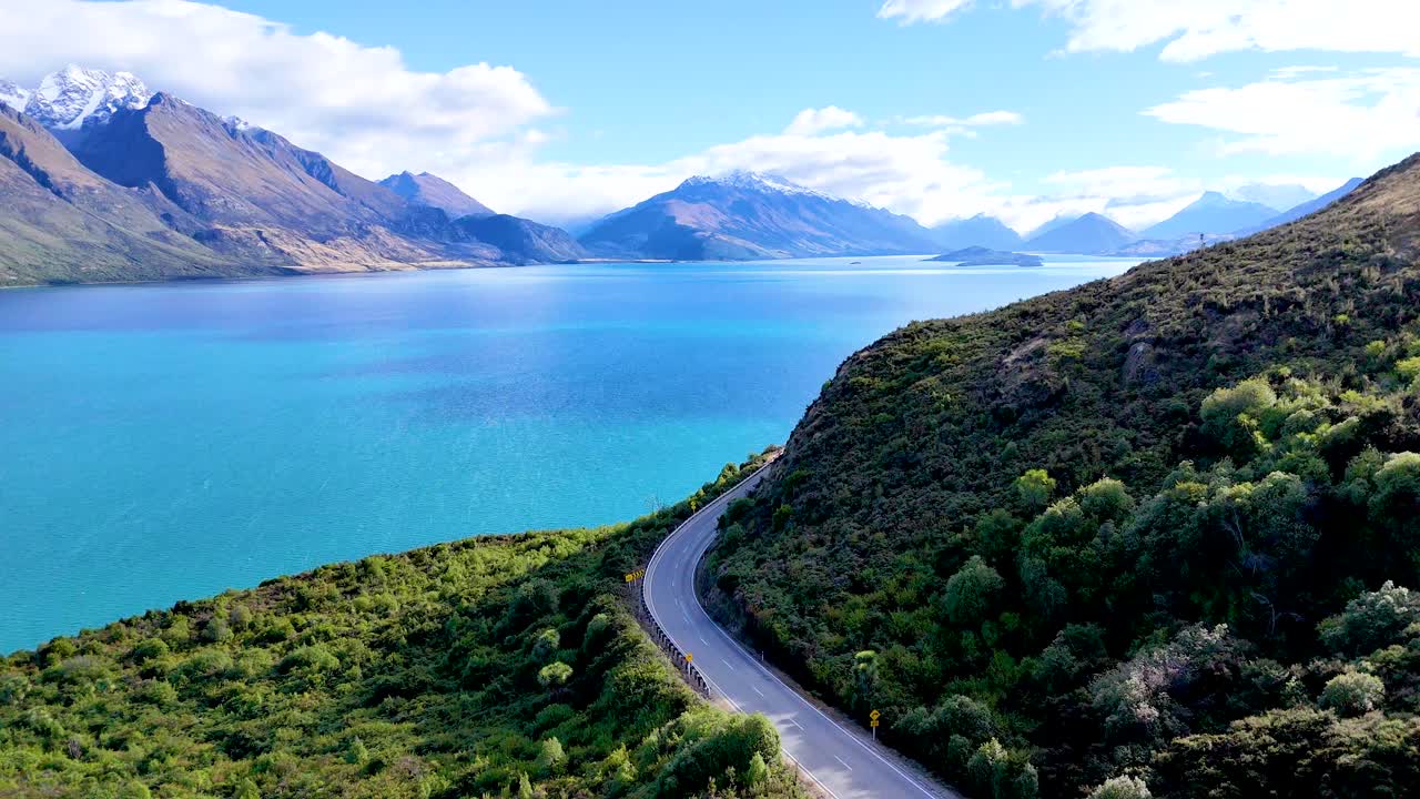 Drone glides above winding road, turquoise lake, and mountains in bright daylight, smooth camera movement