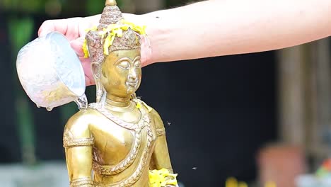 a hand delicately pours water over the head of a golden buddha statue in a serene setting.