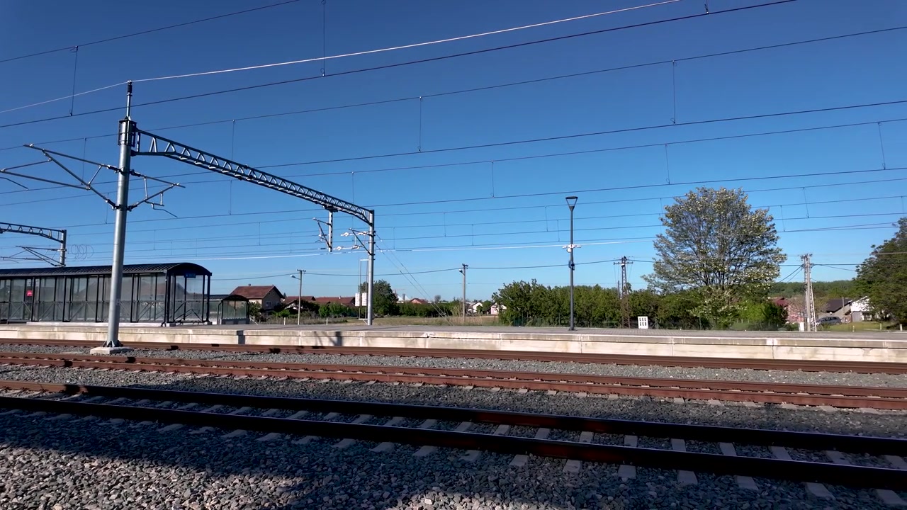 The train station shows empty tracks and an unused platform. The sky is clear, and countryside greenery can be seen in the background. It is a sunny day.